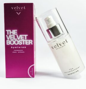 Velvet Skincare Produktfoto von ‚The Velvet Booster Hyaluron‘ mit violetter Verpackung und elegantem Flakon, klar inszeniert auf hellem Hintergrund.