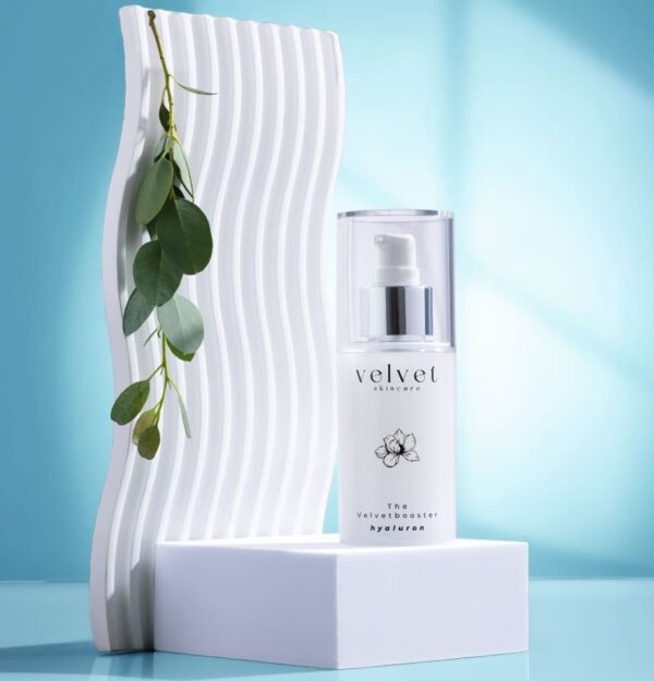 Velvetbooster Hyaluronserum mit Eukalyptusblatt auf weißem Podest vor minimalistischer Kulisse